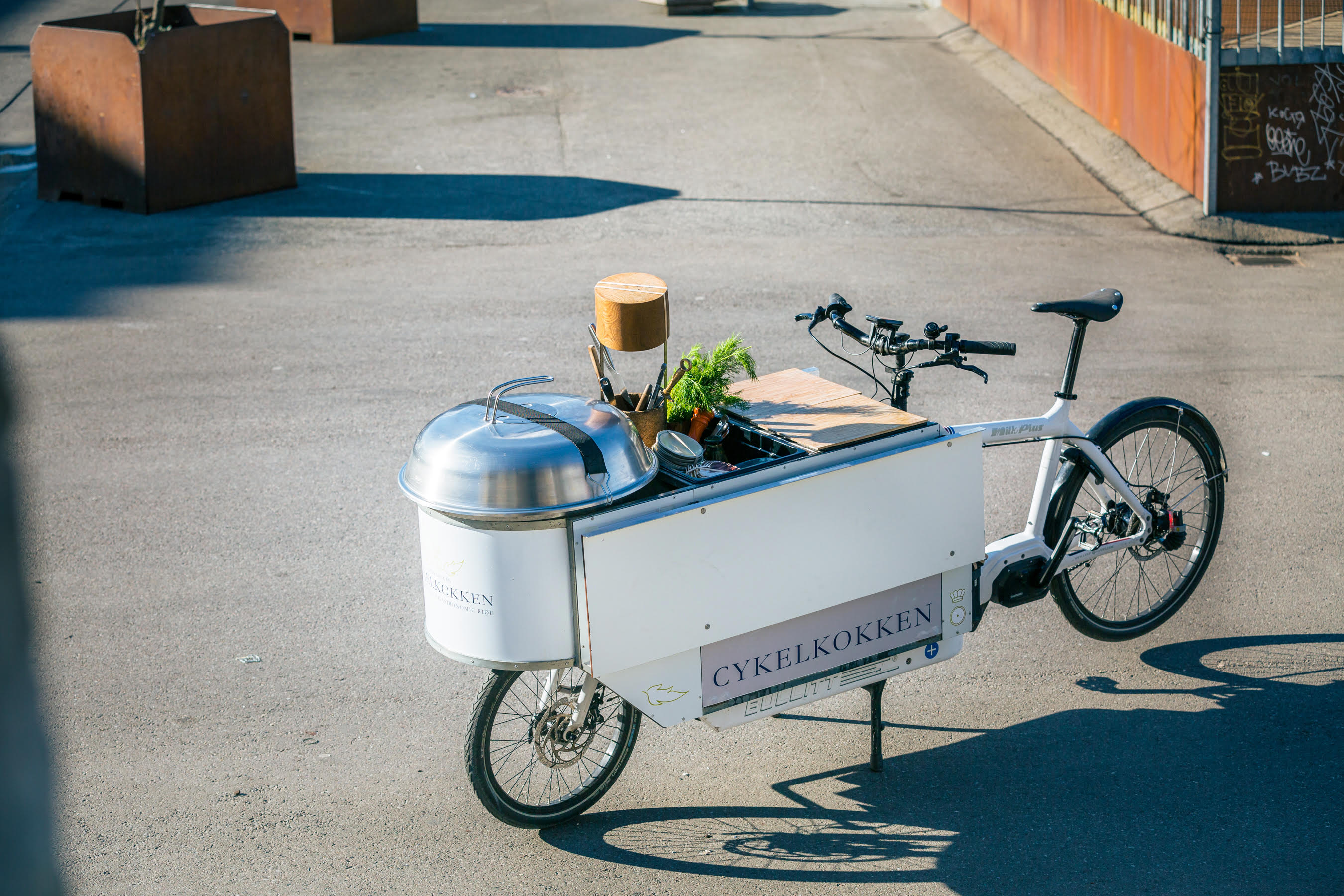A white Bullitt cargo bike with a custom built mobile kitchen mounted on the cargo bay. Cykelkokken logo on the side. Parked in the street and lit by low sunrays.
