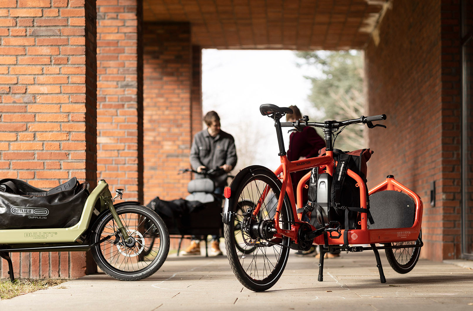 Red electric Bullitt cargo bike and light green Bullitt cargo bike with duffelbag and backpack strapped to the cargo bay, parked in a ray of sun by a red brick building. Two people are standing in the background.
