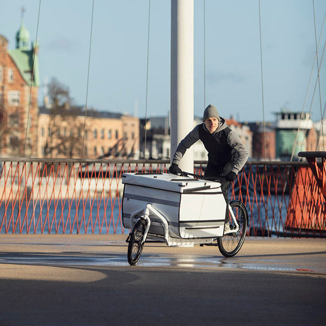 A guy biking in sunny weather on a Bullitt cargo bike with a big white cargo box in the front. He and the Bullitt cargo bike is leaning into a curve. 
