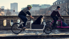 Two men riding Bullitt cargo bikes across a bridge in Copenhagen. One has a dog riding in front.