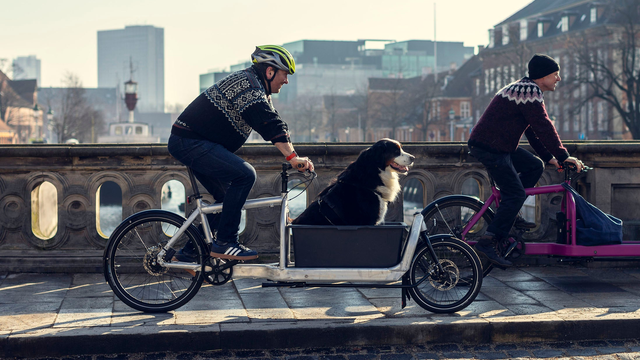 Two men riding Bullitt cargo bikes across a bridge in Copenhagen. One has a dog riding in front.