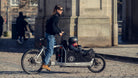 Woman riding a Bullitt cargo bike with her child in the front. She has the wind in her hair and sun on her face and is standing up in the pedals.