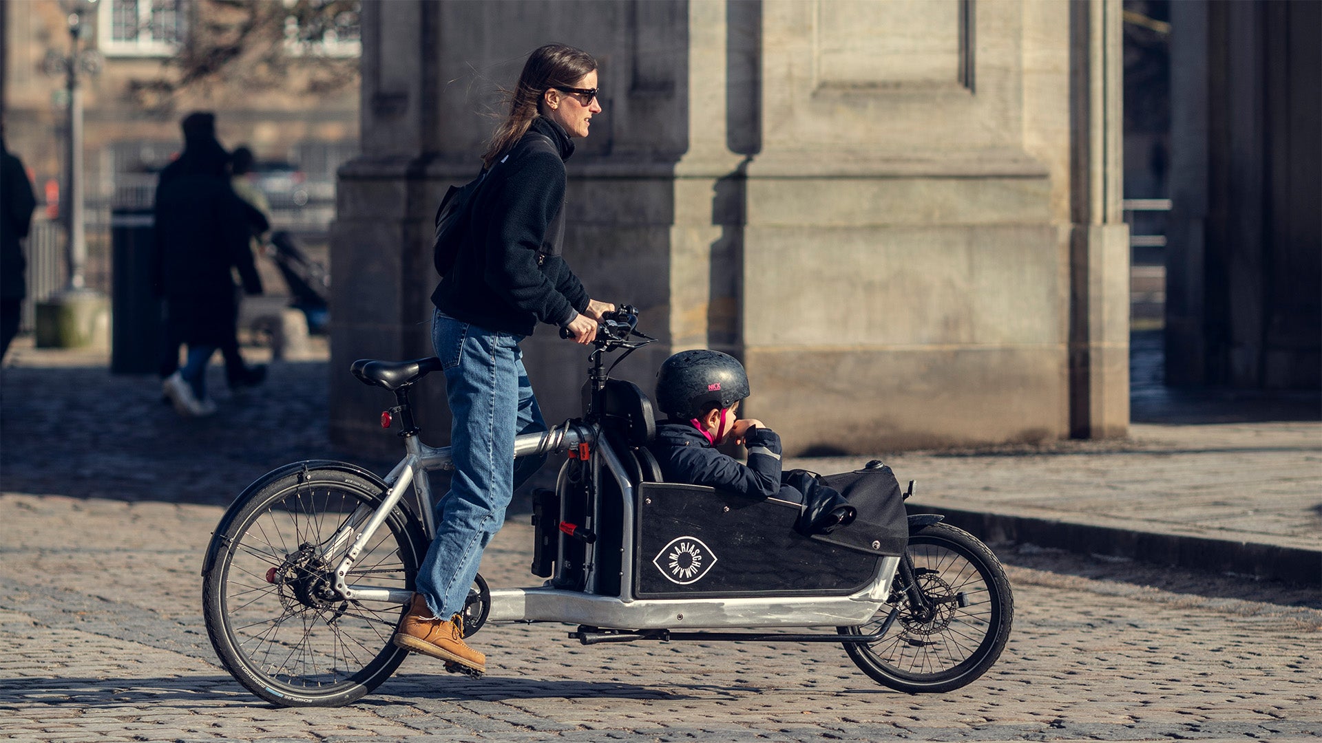 Woman riding a Bullitt cargo bike with her child in the front. She has the wind in her hair and sun on her face and is standing up in the pedals.