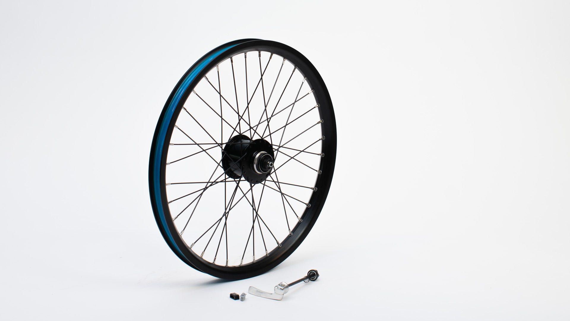 A 20" bike wheel with dynamo hub, without tyre, standing on a white surface. Beside it is an axle with quick release and some smaller parts.