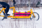 Person riding a yellow Bullitt cargo bike with a pink and yellow bag through a snowy landscape.