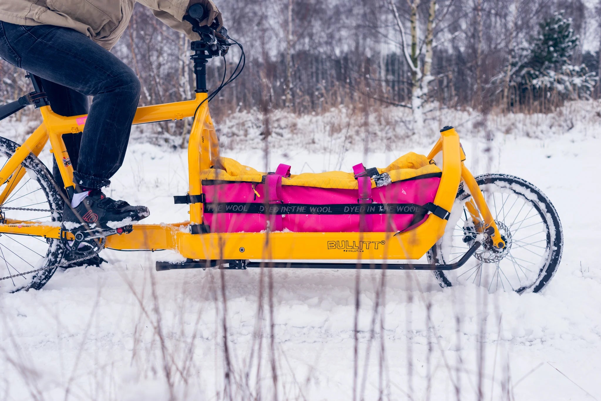Person riding a yellow Bullitt cargo bike with a pink and yellow bag through a snowy landscape.